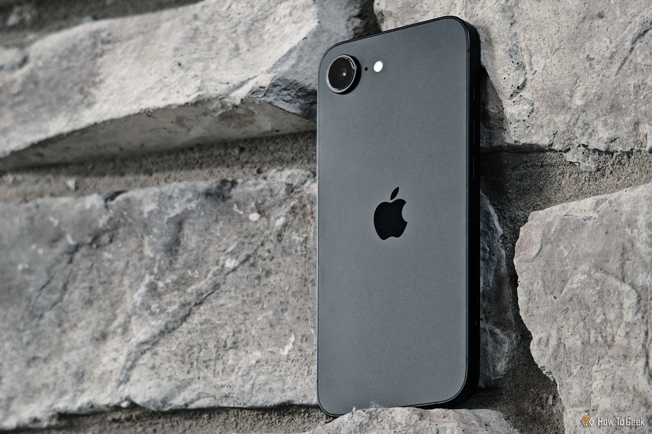Điện thoại iPhone 16e đặt tựa vào bức tường gạch, thể hiện thiết kế và vị trí camera sau.