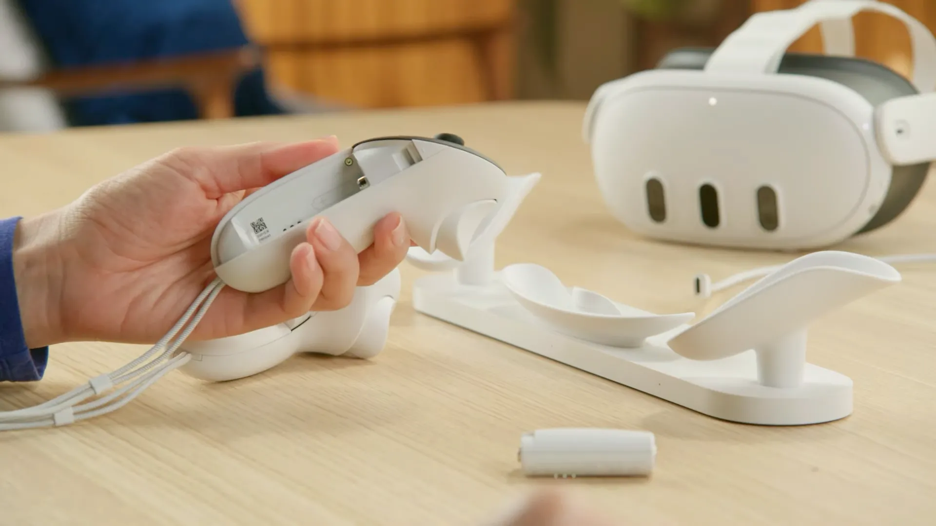 Bàn tay cầm bộ điều khiển Meta Quest 3 Touch Plus với pin sạc, và tai nghe Meta Quest 3 cùng dock sạc trong nền