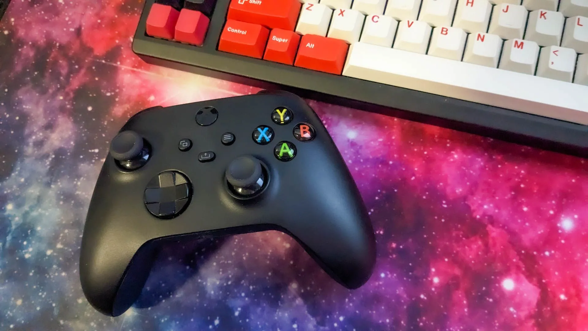 Bộ điều khiển Xbox và bàn phím cơ bên cạnh miếng lót chuột có hình không gian, tượng trưng cho một góc gaming hiện đại và các phụ kiện cần thiết cho game thủ.